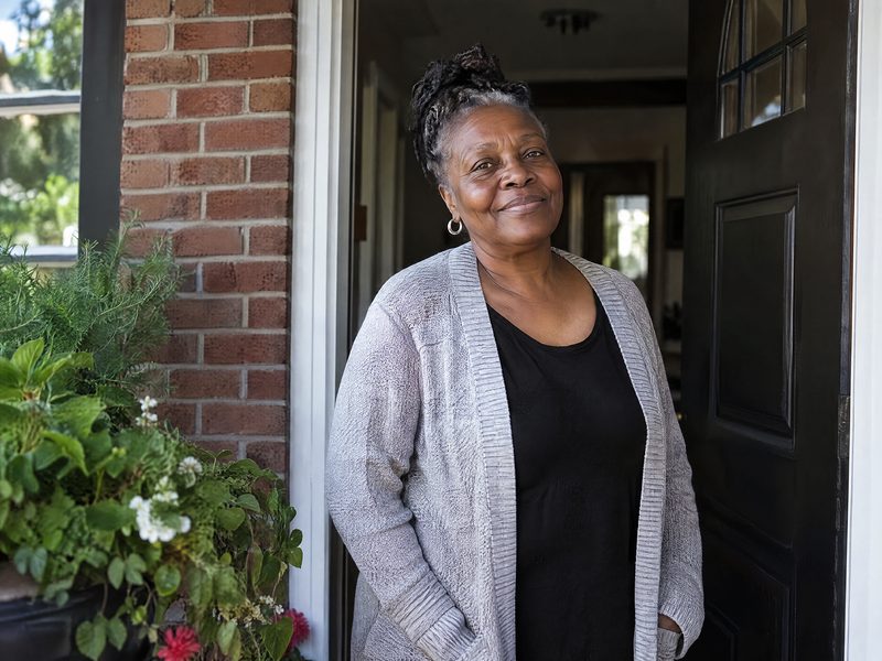 A smiling woman in a gray cardigan standing in the open front door of her brick house